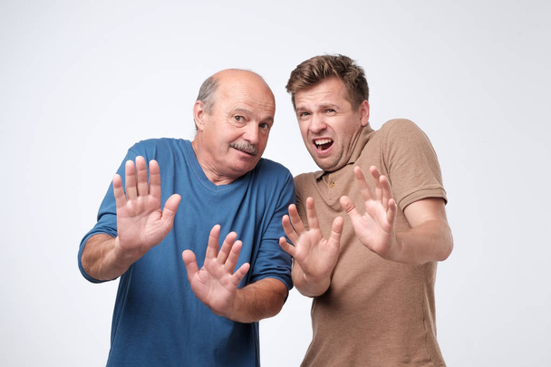 Indoor shot of stunned funny caucasian men tilting backwards and pulling raised palms towards camera, shouting, being shocked and afraid. Mature son and father are shocked with news. - Fotografie, Obrázek