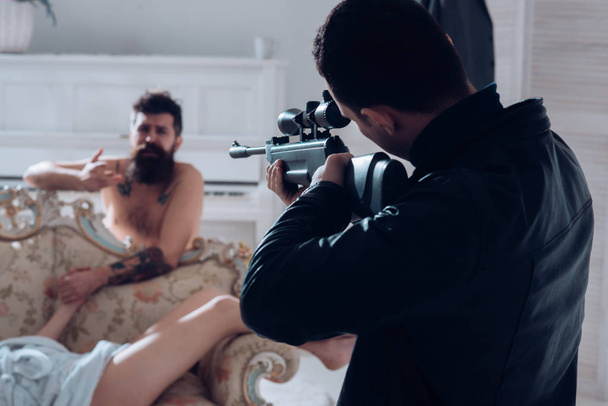 Cheating and jealousy concept. Husband found lovers, killed wife and threatening to bearded lover. Man with beard naked, pray for mercy at gunpoint, interior background. Man at gunpoint of killer - Photo, Image