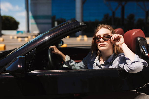 Young beautiful lady in sunglasses thoughtfully looking in camera driving cabriolet car on city streets outdoor - Foto, imagen