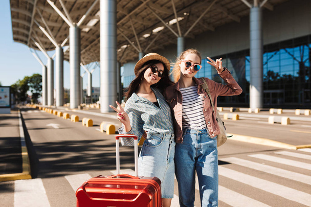 Two beautiful smiling girls in sunglasses happily looking in camera with red suitcase outdoor near airport - Photo, Image