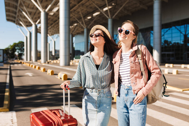 İki gülümseyen güneş gözlüğü sevinçle bir kenara kırmızı çanta ve sırt çantası Havaalanı'nın yakınlarında açık ile seyir kızlarda güzel - Fotoğraf, Görsel