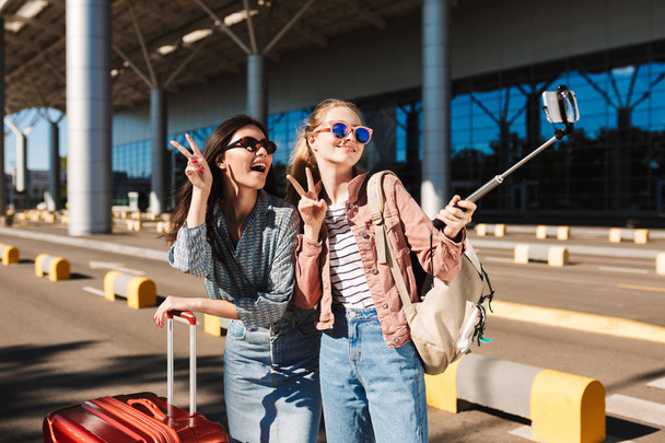 Две веселые девушки в солнечных очках счастливо показывают два пальца жест, делая фото на мобильном телефоне вместе с чемоданом и рюкзаком на плече на открытом воздухе возле аэропорта
 - Фото, изображение