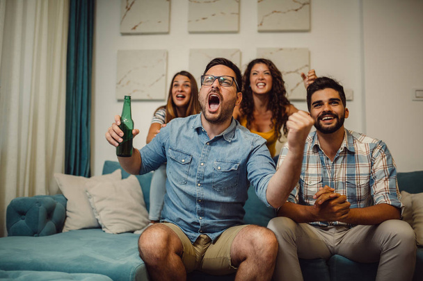 Молодые футбольные фанаты пьют пиво, аплодируют и улыбаются во время просмотра футбольного матча дома
. - Фото, изображение