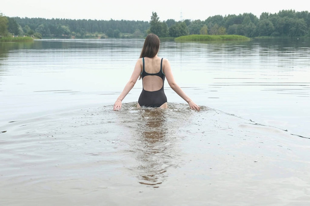 Beautiful brunette woman comes into the water and swim, back view. Beautiful view of the pond in the forest. - Photo, image