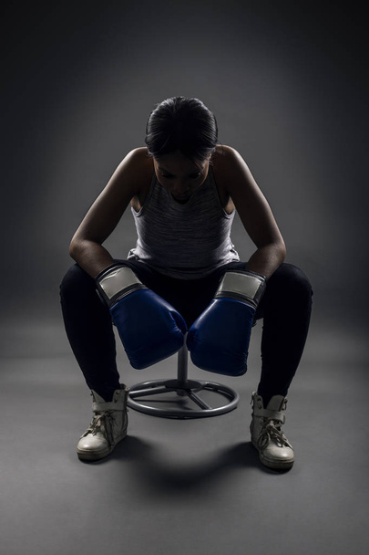 Musta nainen yllään nyrkkeilyhanskat etsivät vihainen kuin nyrkkeilijä, MMA taistelija tai itsepuolustusta kouluttaja istuu treenauksen jälkeen. Hän kuvaa naisten voimaa ja päättäväisyyttä.
.  - Valokuva, kuva