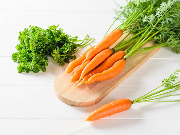 A bunch of carrots and a bunch of parsley on a cutting Board on a white table. The concept of vegetarian food with natural raw products. - Photo, Image