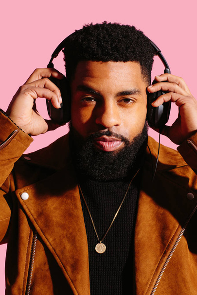Homem afro-americano elegante, bonito e legal com barba, ouvindo música, olhando para a câmera, isolado no fundo do estúdio rosa
 - Foto, Imagem