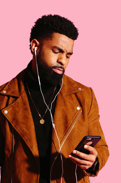 Portrait of a cool man with beard and headphones looking at phone with a frowning reaction, isolated on pink studio background - Photo, image