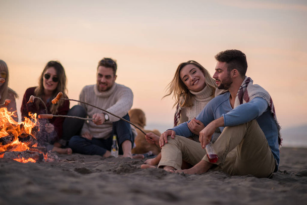Group of young friends sitting by the fire at autumn beach, grilling sausages and drinking beer, talking and having fun - Fotoğraf, Görsel