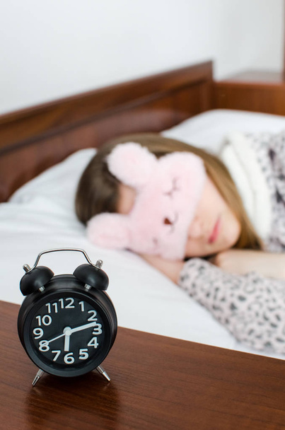 Young woman girl in cute sleep mask and warm pajamas sleep with alarm clock near the bed in the morning. Sleeping girl and alarmclock in bedroom. Teenager afraid to wake up late. Oversleep concept. - Photo, Image