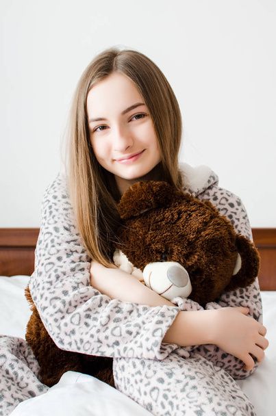 Ευτυχισμένος νεαρή γυναίκα με ξανθά μαλλιά, κάθεται στο κρεβάτι στην κρεβατοκάμαρά της σε μια αγκαλιά με ένα γεμισμένο ζώο παιχνίδι. Αγαπημένο αρκουδάκι καφέ στα χέρια των teen κορίτσι σε ζεστά πιτζάμες. - Φωτογραφία, εικόνα