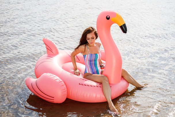Outdoor portrait of beauty brunette model in colorful summer swimwear. Young beautiful woman sitting on pink inflatable mattress in form of flamingo at daytime, wavy water background - Foto, imagen