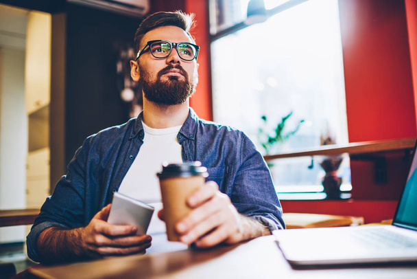 Thoughtful bearded young entrepreneur in casual outfit thinking on developing startup project holding smartphone and coffee to go in hands.Contemplative male blogger with mobile pondering on ideas - Foto, imagen