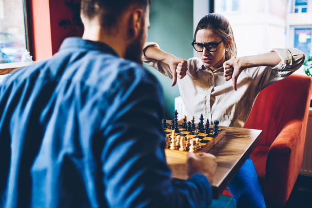 Serious professional female player accepted challenge from male rival and gesturing thumb down showing fail in intelligent game in chess.Angry woman with loser sign deriding opponent during battle - Foto, Bild