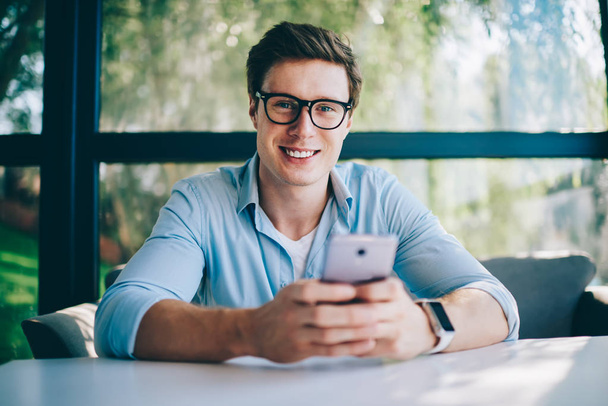 Portrait of cheerful male blogger smiling at camera while messaging in online chat with friend on modern smartphone.Positive hipster guy laughing and installing mobile app on phone using 4G internet - Фото, изображение