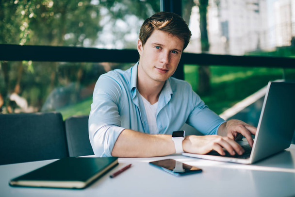 Портрет молодого человека, работающего удаленно за современным ноутбуком и улыбающегося в камеру. Позитивный мужчина графический дизайнер ищет информацию на сайтах на цифровых нетбуках, сидящих в кафе
 - Фото, изображение