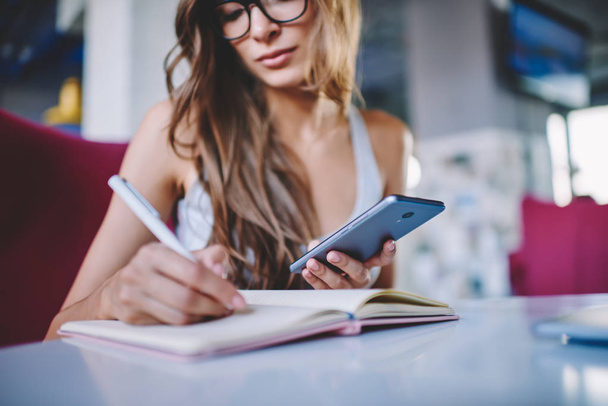 Rajatut näkymä fiksu nuori nainen kirjoittaa ylös tietoja muistilehtiö internet-sivusto tekee kotitehtäviä käyttäen moderni älypuhelin laite ilmainen 4G-yhteys istuu pöydässä kahvilassa
 - Valokuva, kuva