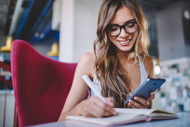 Positive young woman in eyeglasses making notes in notepad sitting at table and using free wifi connection on smartphone for search information.Cheerful student reading notification on cellula - Foto, Bild