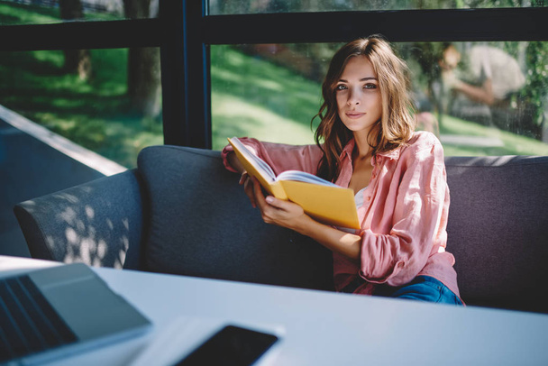 Portrait of intelligent charming young woman dressed in casual wear resting on comfortable couch with literature book in hands in modern cafe interior.Smart hipster girl preparing for upcoming exams - Foto, Imagen
