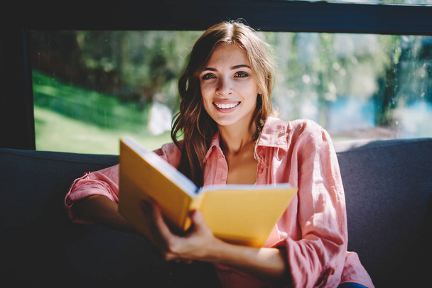 Portrait of happy young woman with literature book in hands spending free time on hobby in cafe interior.Positive hipster student enjoying bestseller resting on cozy couch in coworking space - Foto, Imagen