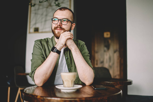 Pondering bearded young man looking away while thinking on plans sitting at coffee table with tasty beverage in cafe.Dreaming hipster guy in eyeglasses enjoying break with aroma drink in coffee shop - 写真・画像