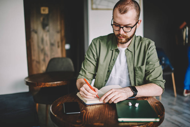 Pensive bearded student in eyeglasses for vision correction writing down text information of studying seminar in notepad sitting at coffee table in cafeteria.Young man making notes in notebook - Fotó, kép