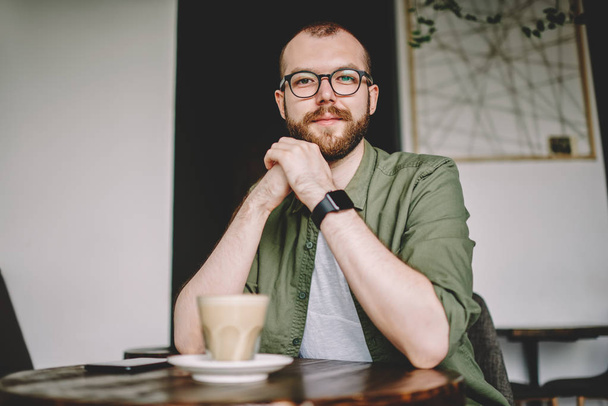 Portrait of bearded hipster guy dressed in casual wear looking at camera while enjoying recreation time in modern cafeteria with tasty coffee drink.Handsome young man in eyeglasses resting in cafe - Φωτογραφία, εικόνα