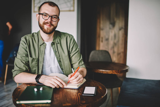 Portrait of successful male blogger in eyewear smiling at camera while writing down notes of publication on website.Positive young man sitting at table with notepad in cafeteria preparing homework - Фото, изображение