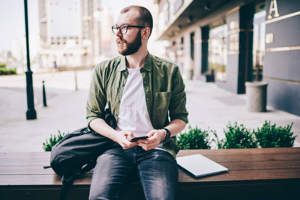 Caucasian young man in eyeglasses looking away while sitting outdoors and holding modern smartphone device in hands.Pondering hipster guy installing app on mobile phone using 4G internet connection - Photo, Image