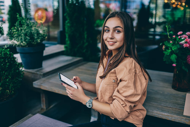 Portrait of cheerful brunette woman holding mobile phone for chatting and blogging on free time, pretty smiling hipster girl looking at camera connecting smartphone to wireless internet at cafe - Fotó, kép