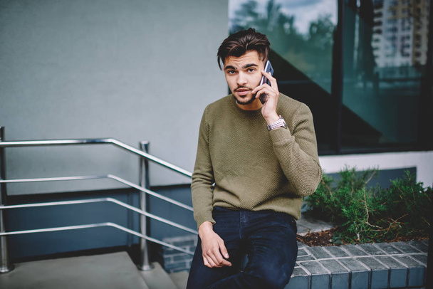 Portrait of serious bearded man having mobile phone conversation with service operator, handsome hipster guy sitting outdoors making telephone call in roaming for communication via telephone - Valokuva, kuva