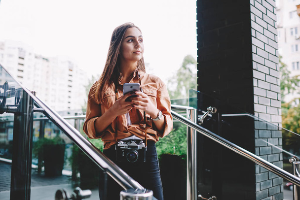 Pensive hipster girl looking away holding mobile phone connected to 4G internet standing outdoors with vintage camera, woman amateur using application for networking  standing on urban setting - Foto, Imagem