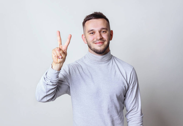 Closeup portrait of happy, excited successful young man giving peace, victory or two sign on gray background. Positive emotions, face expressions, feelings, attitude, reaction, perception - Photo, Image