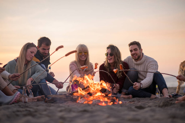 Group of young friends sitting by the fire at autumn beach, grilling sausages and drinking beer, talking and having fun - Foto, Imagem