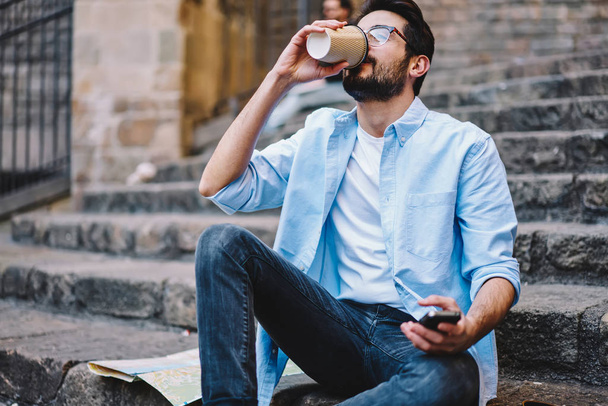 Good looking hipster guy in casual wear enjoying coffee to go outdoors resting in town, young man drinking hot aroma beverage from eco cardboard cup in morning sitting on stairs on spring weekends - Fotoğraf, Görsel