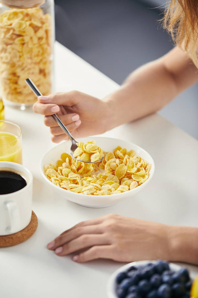 vista parcial da mulher com colher sentada à mesa com flocos de milho e xícara de café no café da manhã
 - Foto, Imagem