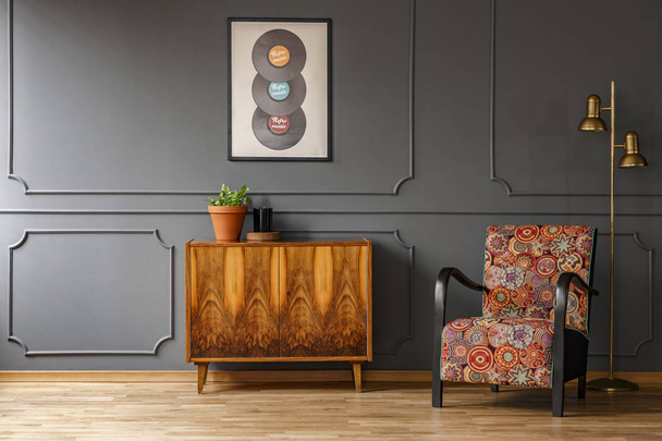 Real photo of wooden retro cupboard with fresh plant and candle standing in dark grey living room interior with colorful armchair, gold lamp and poster with vinyls - Valokuva, kuva