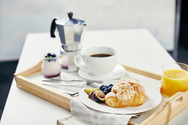 vista de cerca de croissant con arándanos frescos y trozos de ciruela, vaso de jugo, taza de café y yogur para el desayuno en bandeja de madera sobre mesa blanca
 - Foto, Imagen