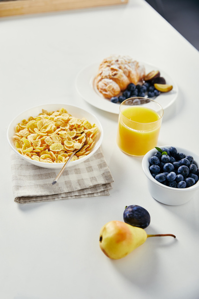 Close-up van cornflakes in bowl, glas sap en croissant met bosbessen en stukjes pruimen voor het ontbijt op wit tafelblad - Foto, afbeelding