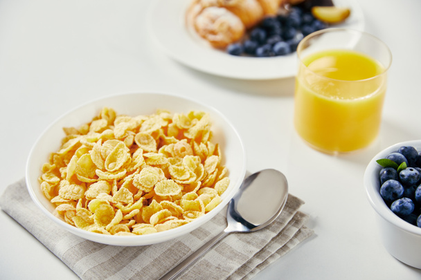 close up view of corn flakes in bowl, glass of juice and croissant with blueberries and plum pieces for breakfast on white tabletop - Fotoğraf, Görsel