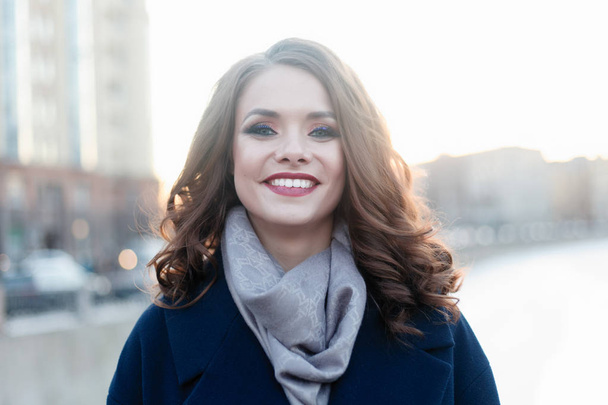 Enthusiastic smiling girl with shiny curls posing in the city in sunlight. Close-up outdoor portrait of enchanting lady in trendy autumn trench with wonderful smile and big blue eyes. - 写真・画像