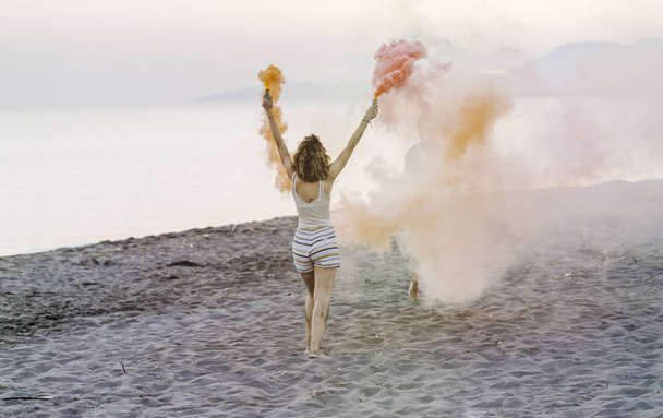 Back view of woman holding smoke bombs and dancing on beach party. - Zdjęcie, obraz