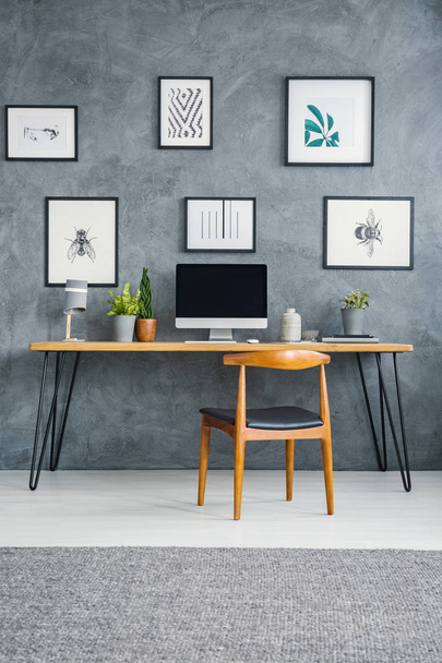 Wooden chair at desk with desktop computer against grey wall with posters in workspace interior. Real photo - Photo, Image