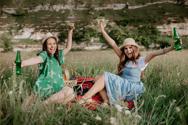 Two attractive young women sitting down on the picnic blanket and have fun. Beer and sandwiches. - Foto, immagini