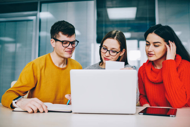 Successful hipster student rewriting information from business card in search job.Positive young people dressed in casual wear collaborating at laptop computer in office sitting at modern laptop - 写真・画像