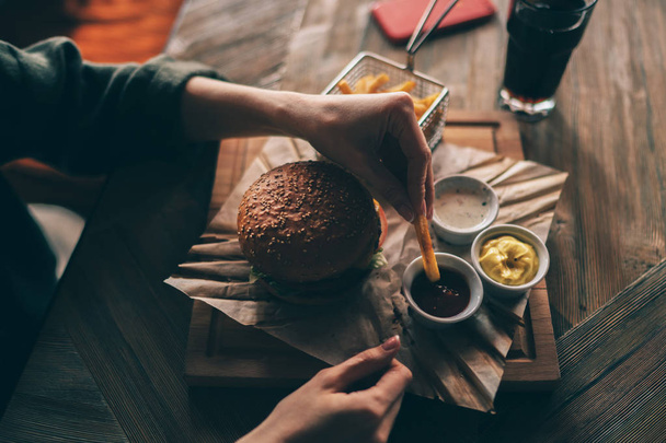 Woman holding cheeseburger and refreshing drink. Young girl holding in hands fast food burger, american meal. Beef burger in hands with french fries on rustic wooden background. Selective focus. - Φωτογραφία, εικόνα