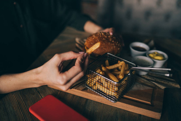 Woman holds burger with hands and fries on the background in cafe. Fresh burger cooked at barbecue in craft paper. American food. Big hamburger with meat and vegetables closeup unfocused at background - 写真・画像