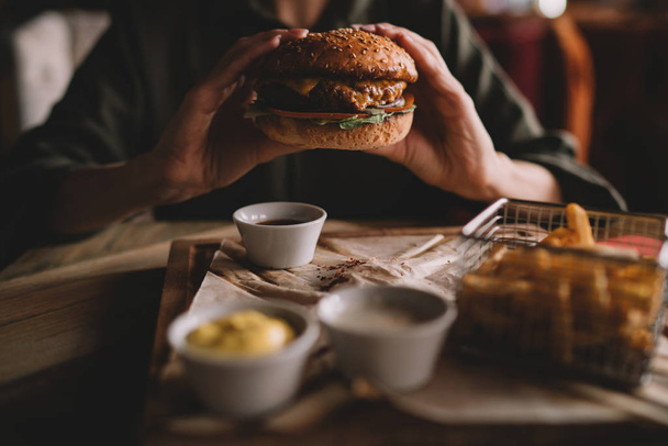 Woman holds burger with hands and fries on the background in cafe. Fresh burger cooked at barbecue in craft paper. American food. Big hamburger with meat and vegetables closeup unfocused at background - 写真・画像
