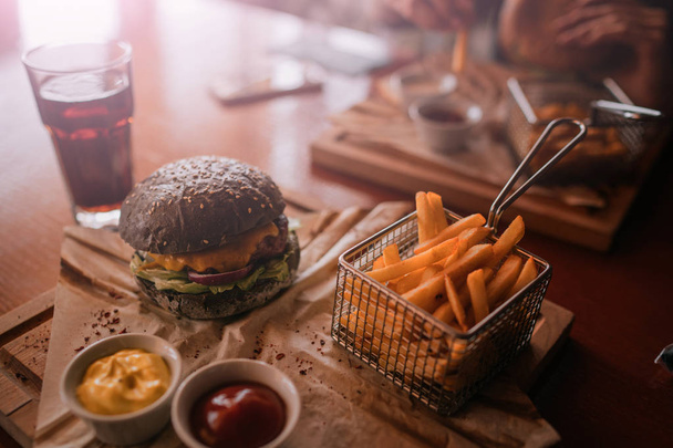 Tasty burger and sauce on wooden tray. Woman eating burger and chips in cafe. People and eating concept. Hamburger, french fries, ketchup, mustard on a wooden board. Toned image. - 写真・画像