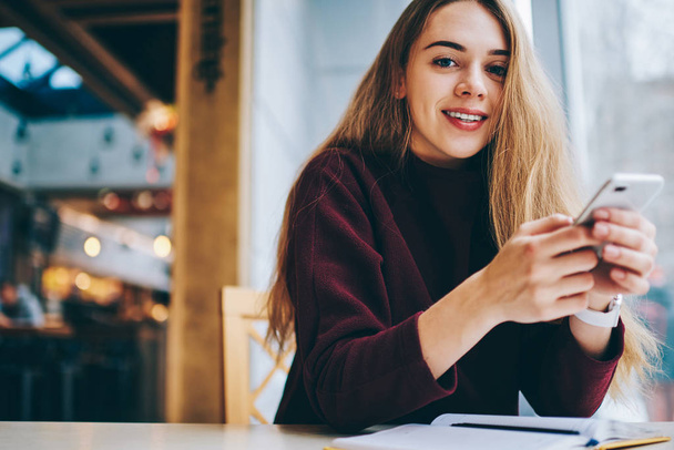 Portrait of positive hipster girl looking at camera and messaging with friends online via application at university campus, young successful woman using modern technology for chatting indoors - Photo, Image